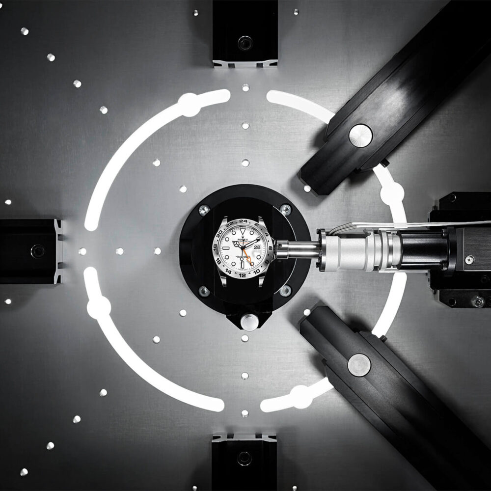 A luxury watch undergoing precise inspection or assembly by robotic arms within a high-tech, illuminated workstation.