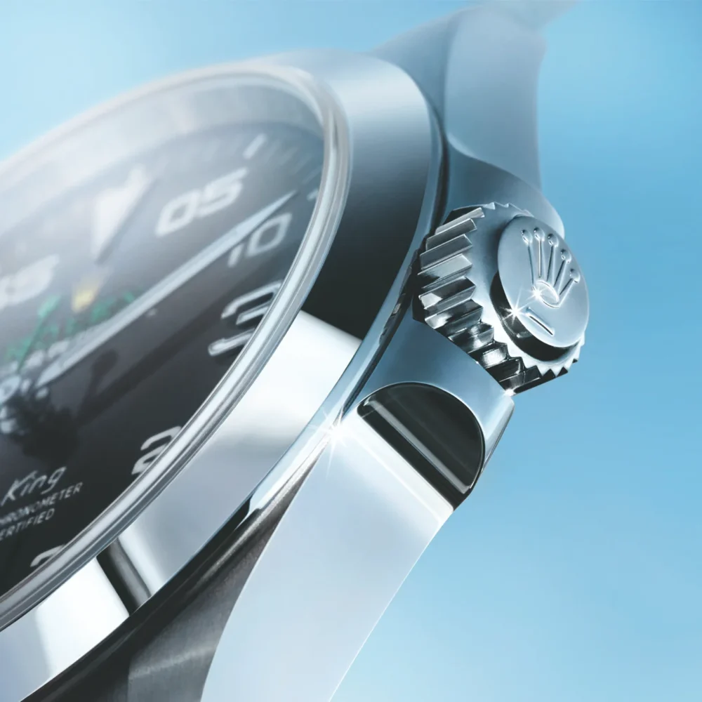 Close-up of a luxury watch showing a detailed view of the crown, bezel, and part of the strap against a light blue background.