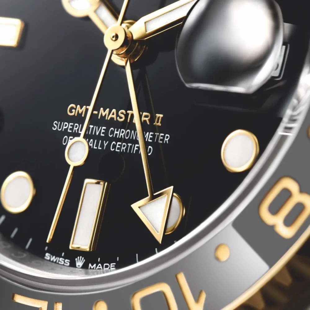 Close-up of a luxury watch face showing the GMT-Master II inscription, gold hands, and hour markers on a black and gray background.