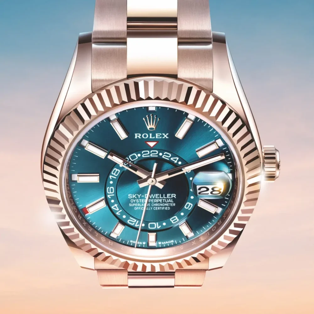 A Rolex Sky-Dweller watch with a blue dial, date window, and dual time zone display against a soft pink and blue gradient background.