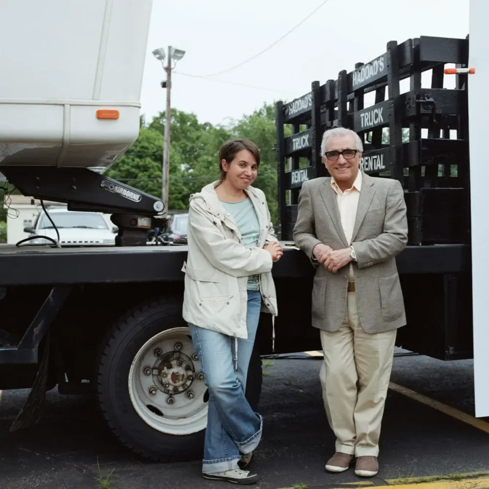A young woman and an older man smiling and shaking hands in front of a semi truck and a black industrial equipment.
