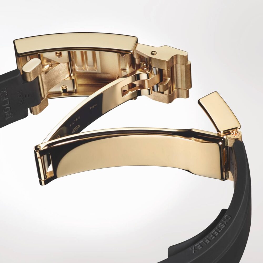 A close-up image of a luxurious gold watch clasp and band against a light background.