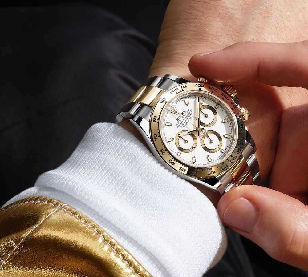 A person adjusts a luxurious gold and silver watch on their wrist, which features a white sleeve and a golden fabric backdrop.