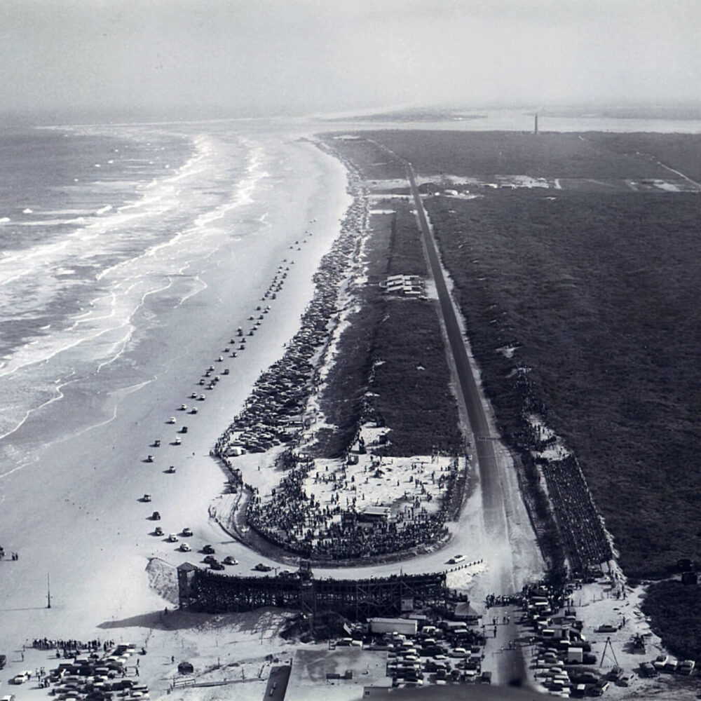 Aerial black and white historic photo of a crowded beach with cars parked along a narrow peninsula.