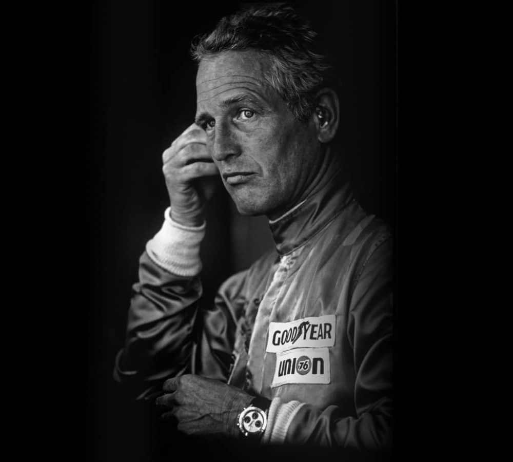Black and white portrait of a mature man in a racing suit with sponsor logos, touching his temple thoughtfully.