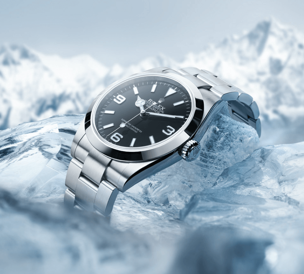 A luxury wristwatch with a metallic strap resting on icy terrain, against a backdrop of snowy mountains.