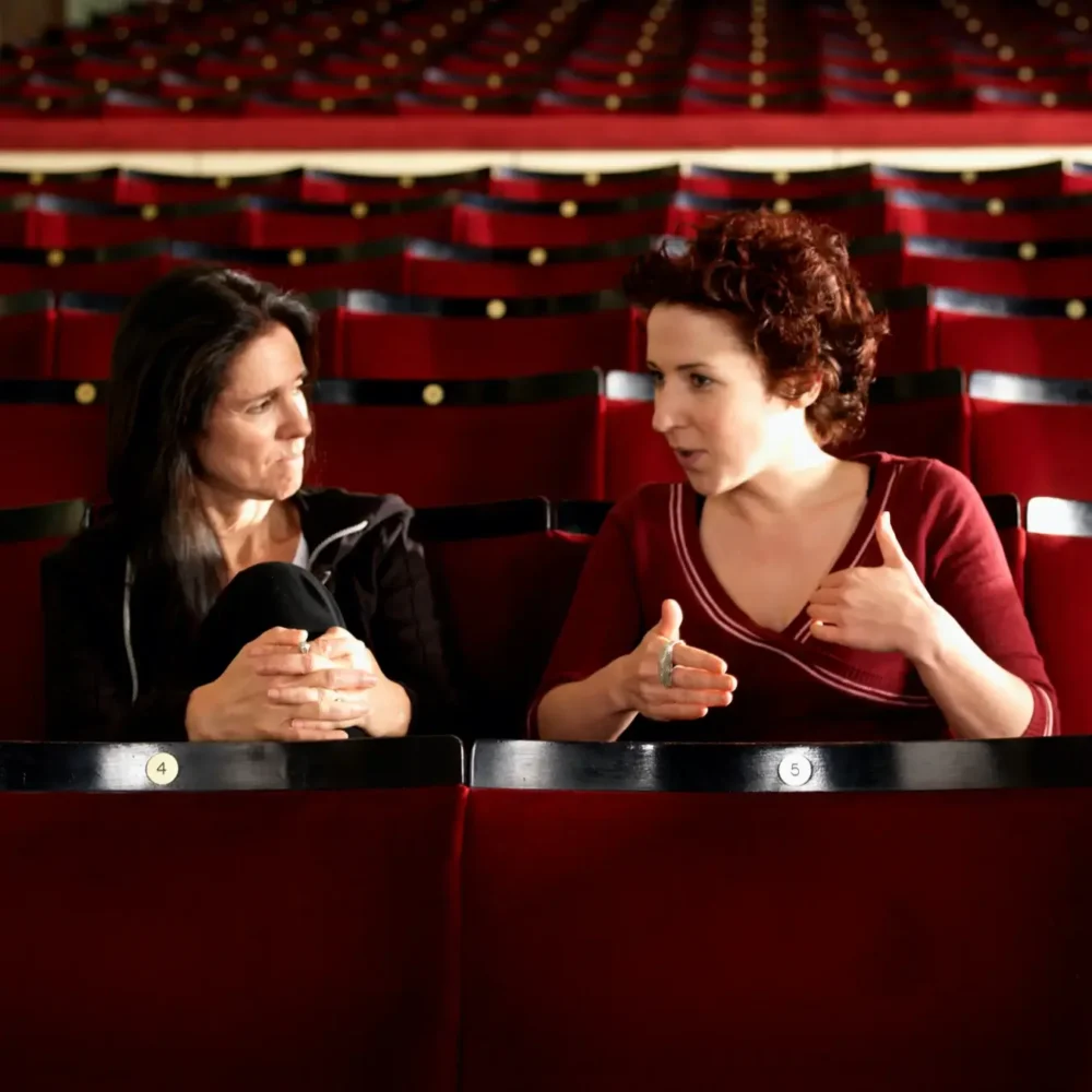Two women conversing in a theater, one gesturing animatedly, surrounded by rows of empty red seats.