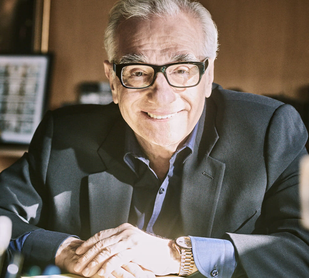 Senior man with glasses smiling at the camera, seated at a desk with papers, in a warmly lit office.