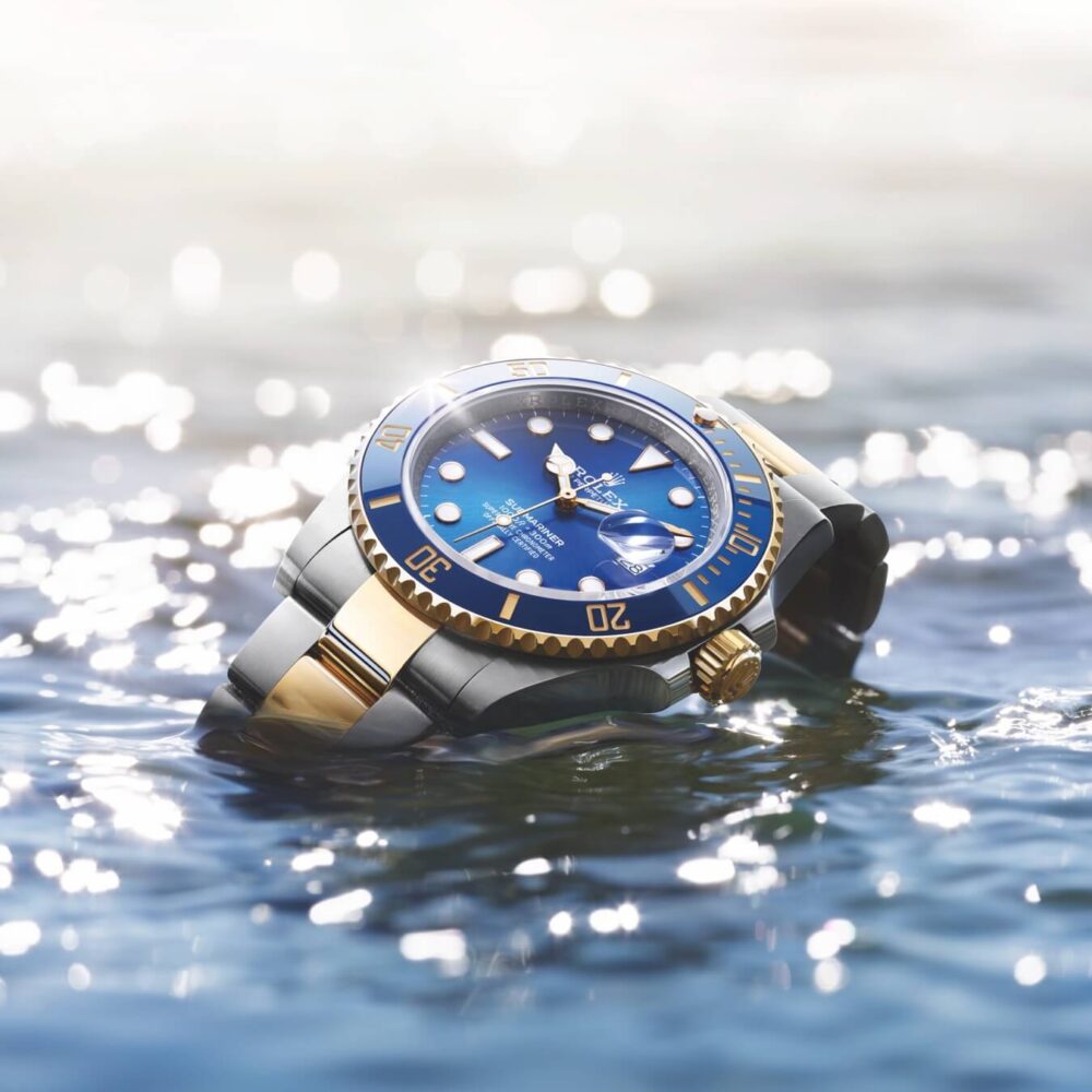 A luxury gold and blue watch partially submerged in shimmering water, reflective light creating a sparkling effect around it.