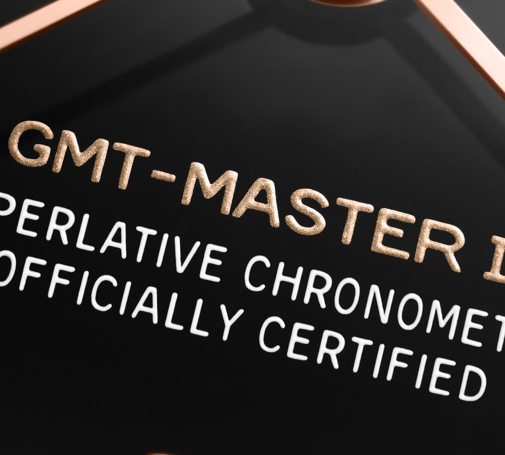 Close-up of a watch dial labeled "GMT-MASTER II SUPERLATIVE CHRONOMETER OFFICIALLY CERTIFIED" with gold lettering on a black background.