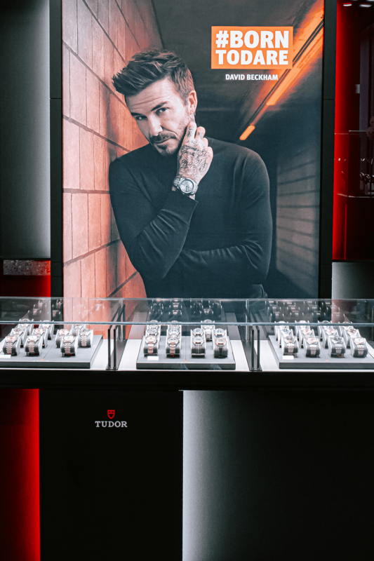 Advertisement featuring David Beckham with a #BORNTODARE slogan above, displayed over a collection of watches in a store.