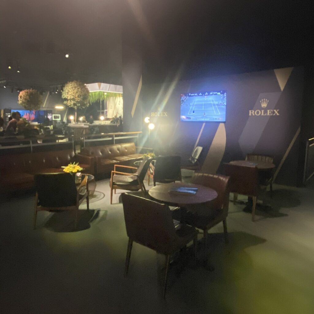 Dimly lit lounge area with leather chairs, tables, and a large screen displaying a tennis match, adorned with Rolex branding.