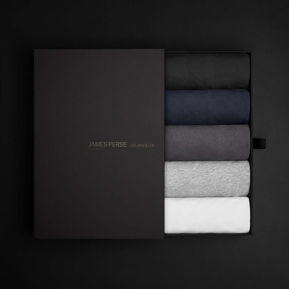 Elegant black box containing neatly stacked James Perse t-shirts in shades of black, navy, blue, gray, and white.