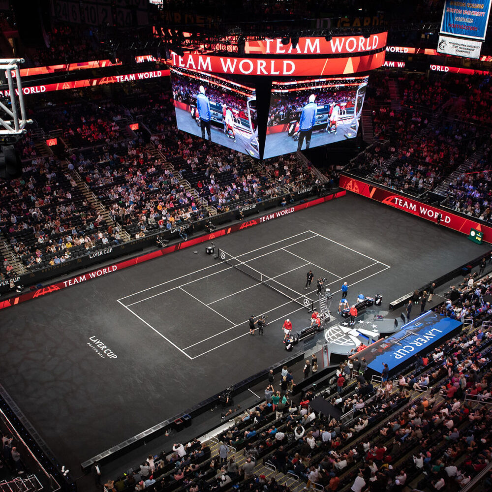 Aerial view of a tennis match at a crowded indoor arena with a large jumbotron displaying the match.