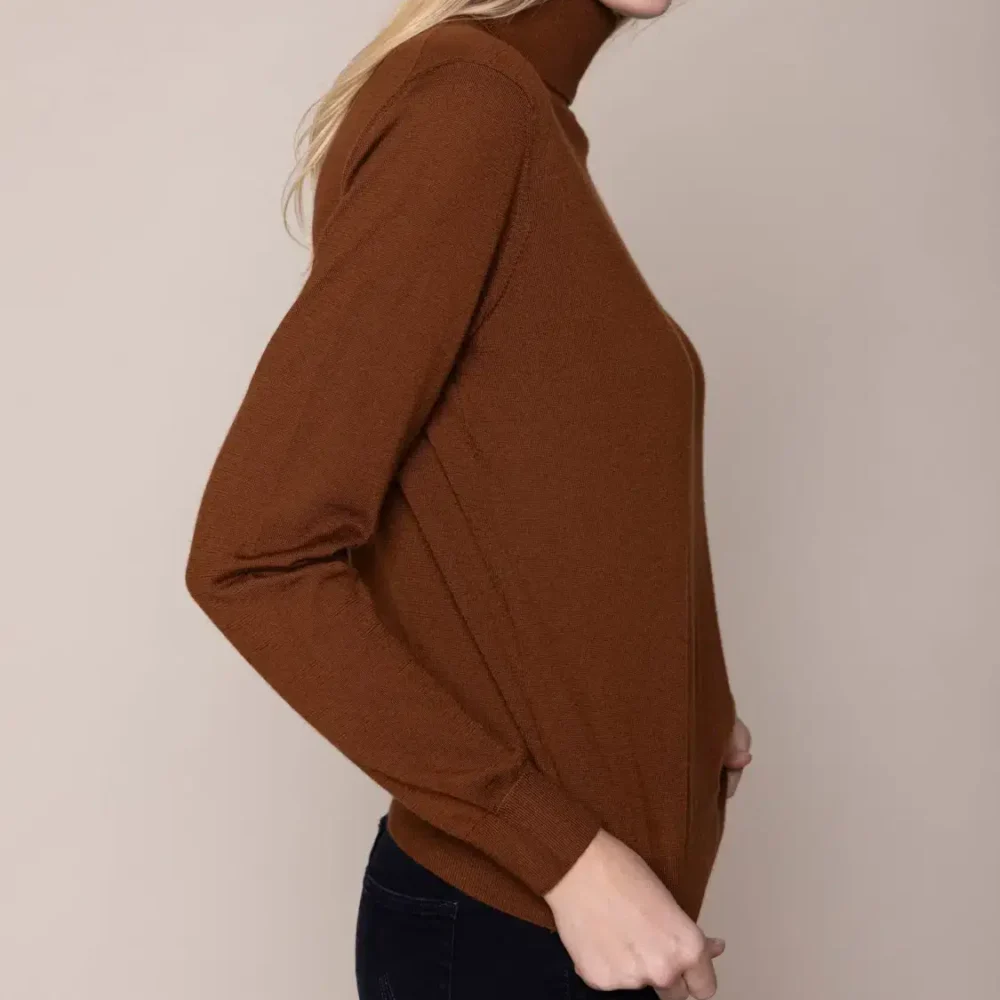 A woman with blonde hair, smiling, wearing a brown sweater and jeans, standing sideways against a beige background.