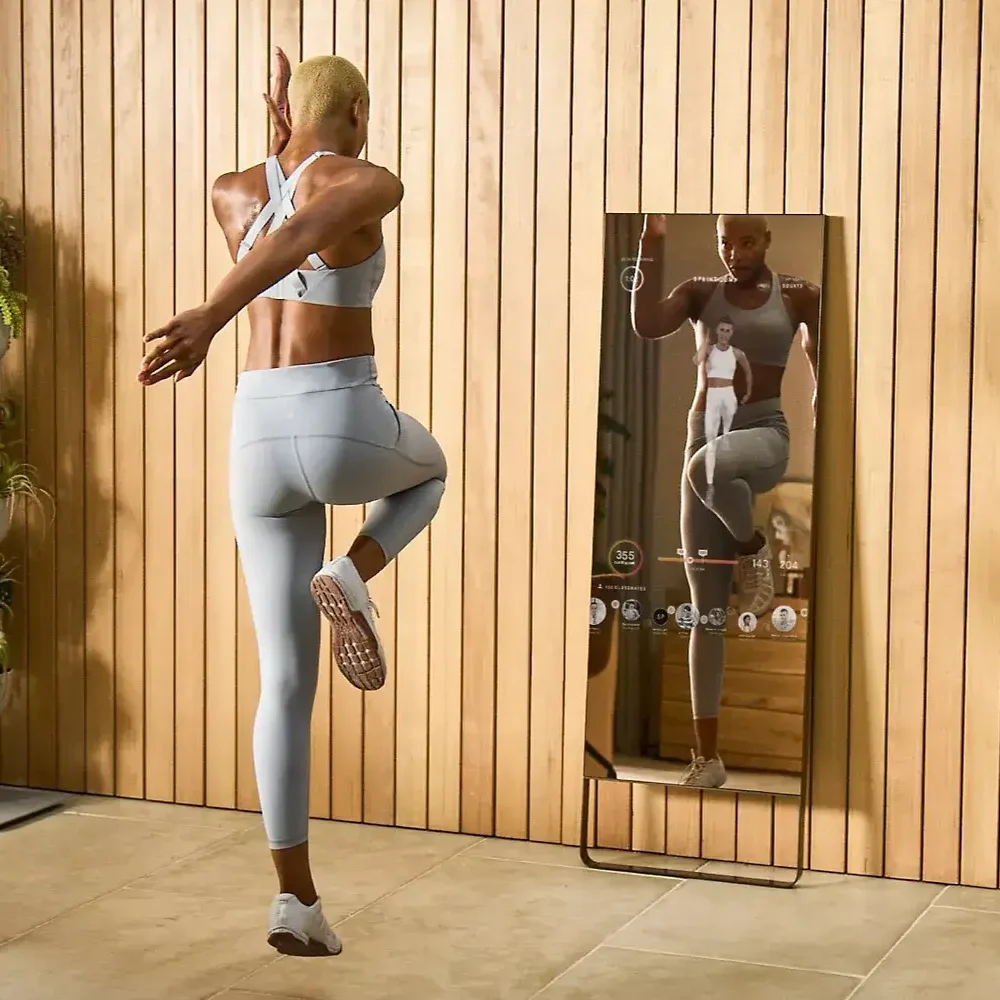 A woman in sportswear exercising in front of a smart mirror that displays her form and workout stats in a room with wooden walls and plants.