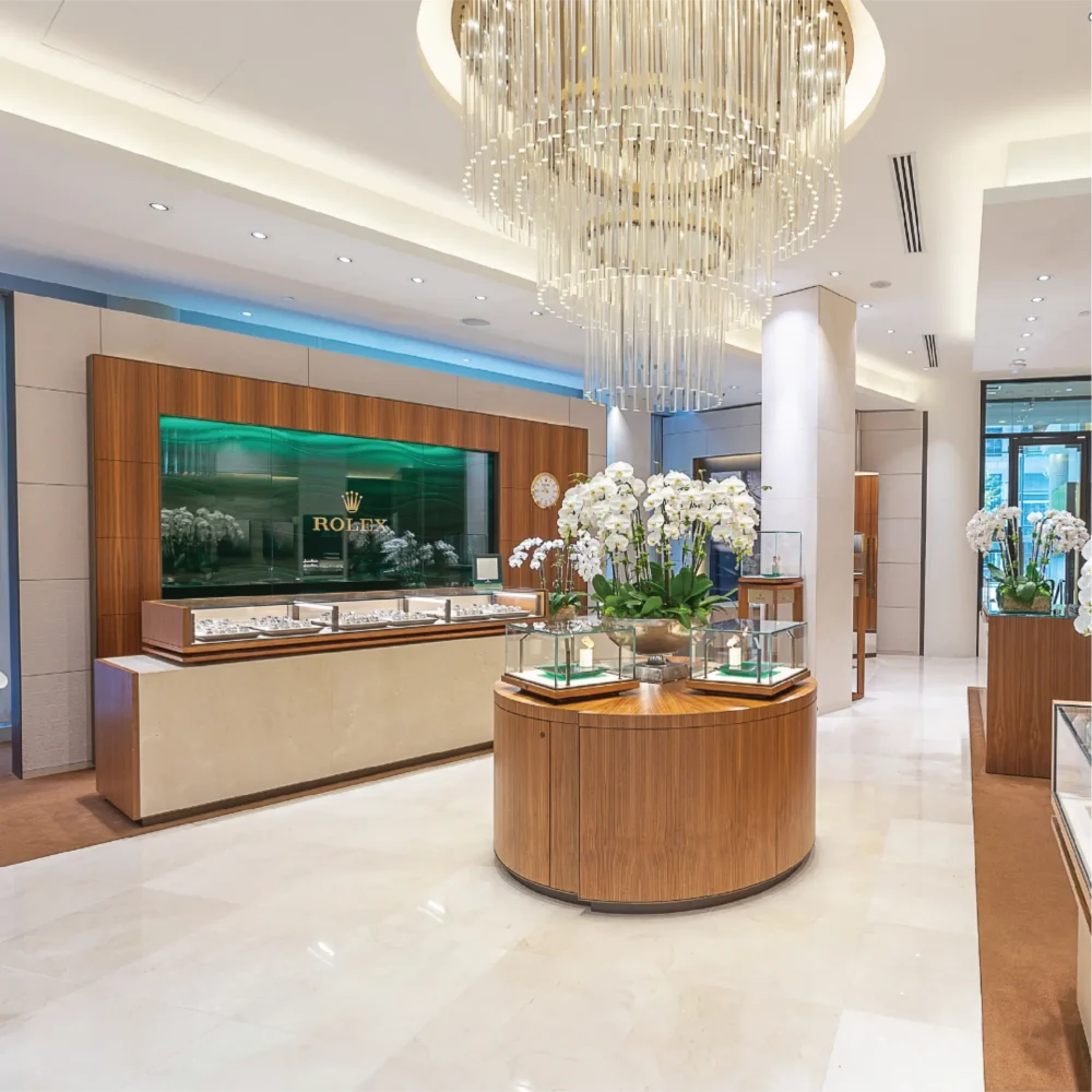 Luxurious watch store interior featuring a Rolex branded counter, glass displays with watches, orchid arrangements, and a striking vertical chandelier.