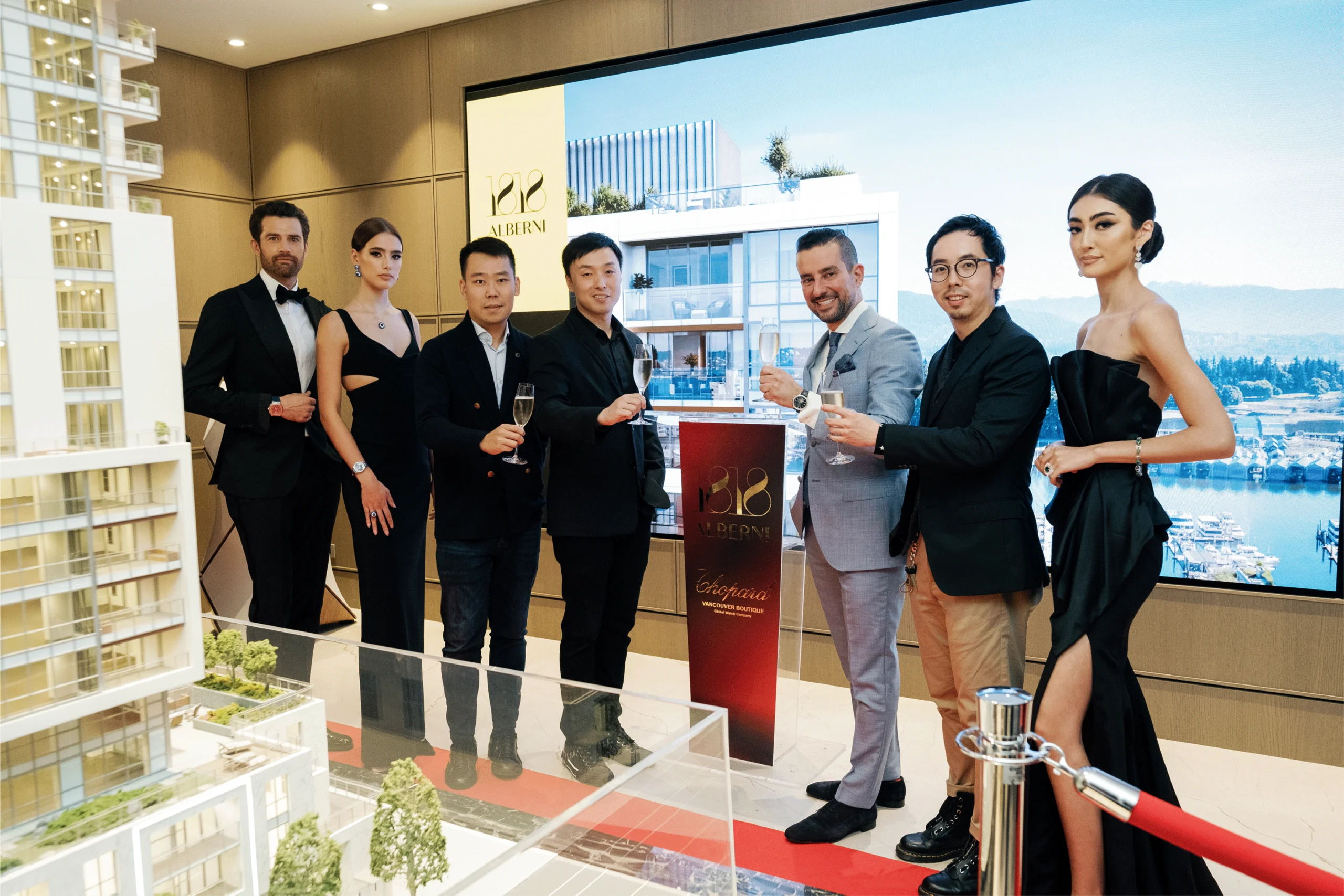 Group of seven people, dressed in formal attire, standing around a model building display at a real estate event.
