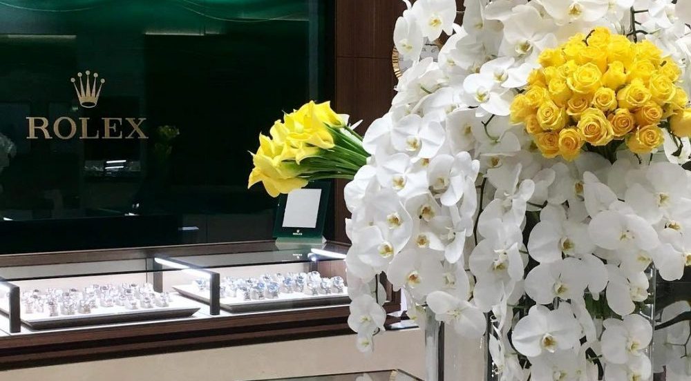 Luxurious Rolex store interior with a foreground of a vibrant floral display featuring yellow roses and white orchids near displays of watches.