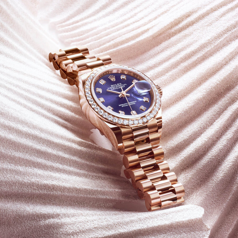 A luxurious rose gold watch with a blue face and diamond-studded bezel, displayed on a textured satin fabric.