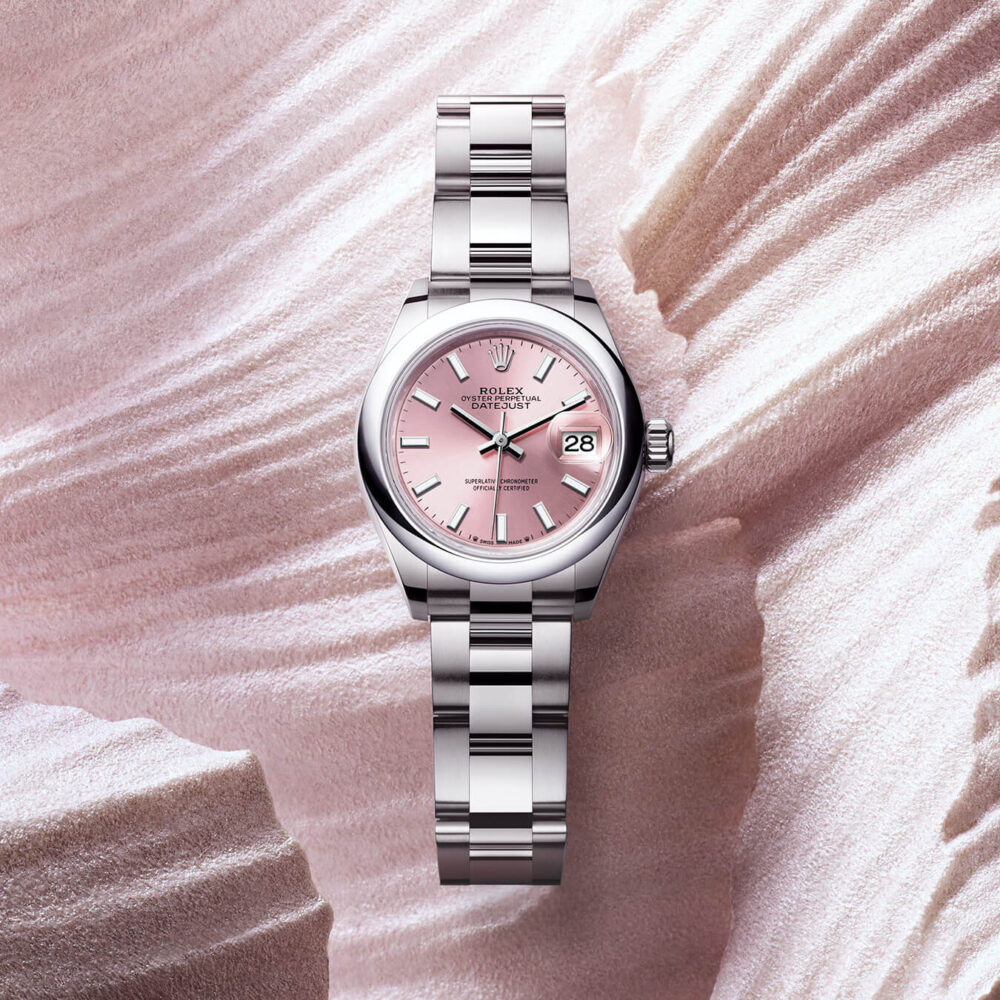 A Rolex watch with a pink dial and silver bracelet set against a textured pink background.