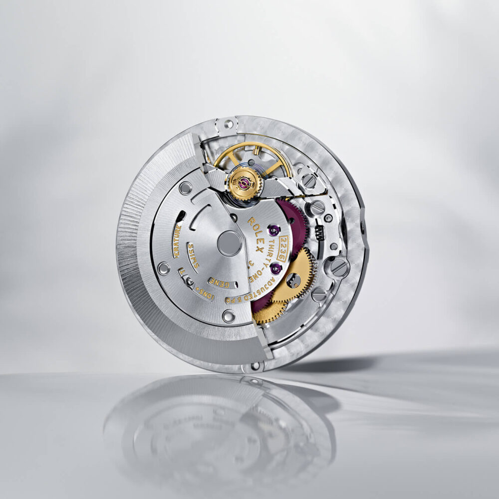 A detailed image of a watch movement with visible gears and components, displayed against a reflective grey background.