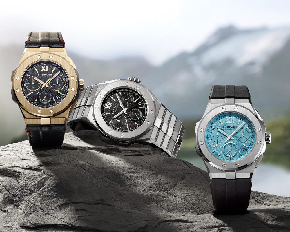 Three luxury watches on a rock with a mountainous backdrop: one gold with a black strap, one silver with a metal band, and one black with a turquoise face and strap.