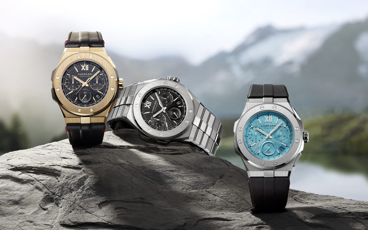 Three luxury watches on a rock with a mountainous backdrop: one gold with a black strap, one silver with a metal band, and one black with a turquoise face and strap.