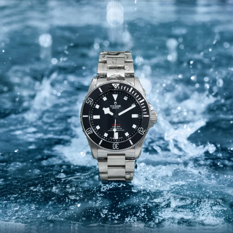 A stainless steel wristwatch with a black dial, red seconds hand, and luminous markers is surrounded by splashing water droplets.
