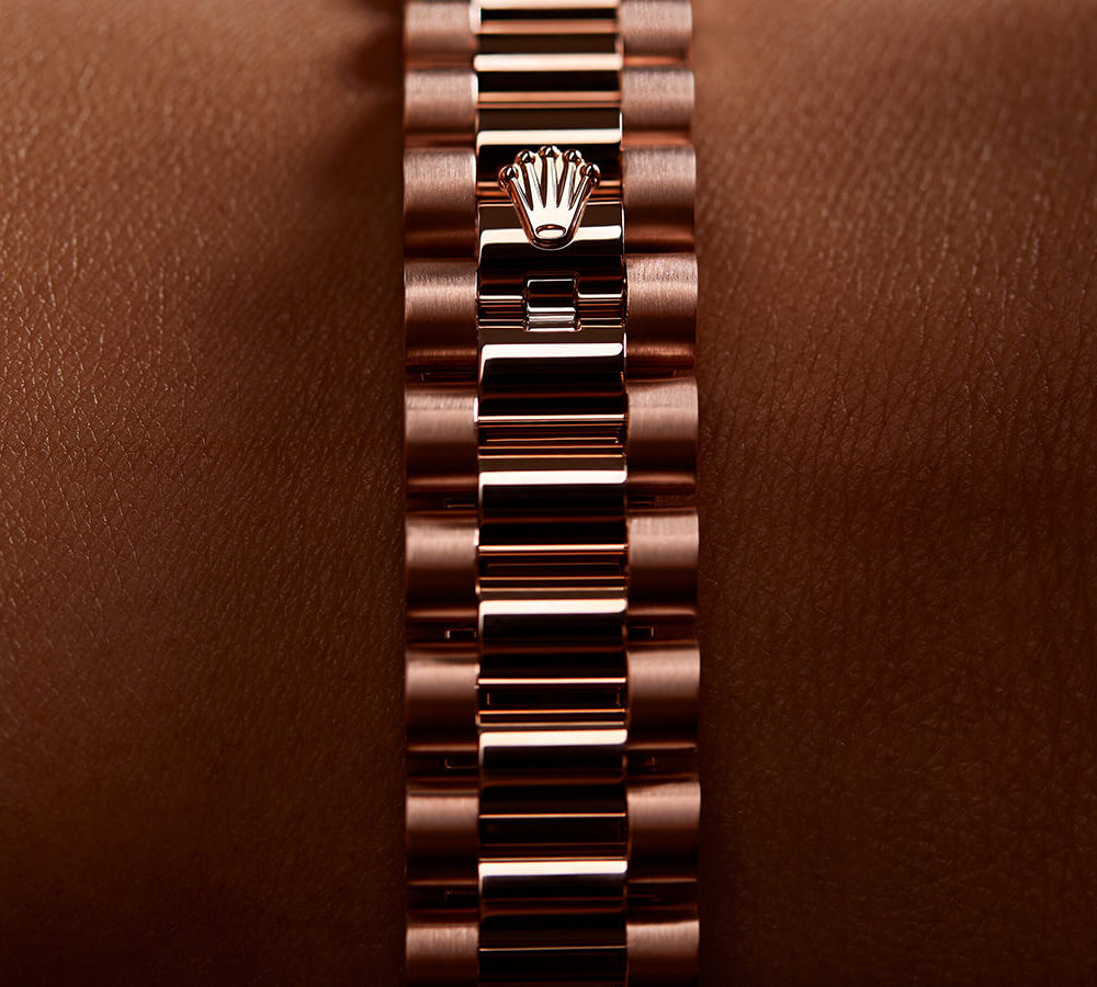 Close-up of a rose gold watch bracelet with a branded clasp, set against a smooth brown background.