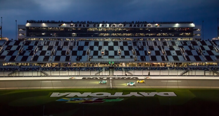 Nighttime view of a race at Daytona International Speedway with illuminated grandstands and several cars on track.