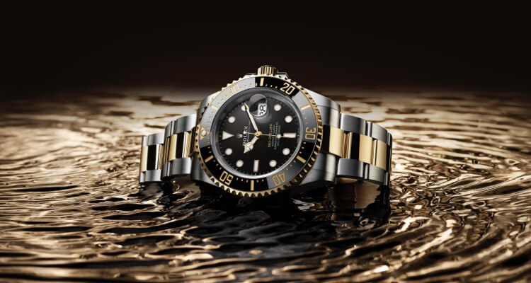 A luxury diver's watch with a black and gold bezel on a two-tone metal bracelet, partially submerged in water with ripples.