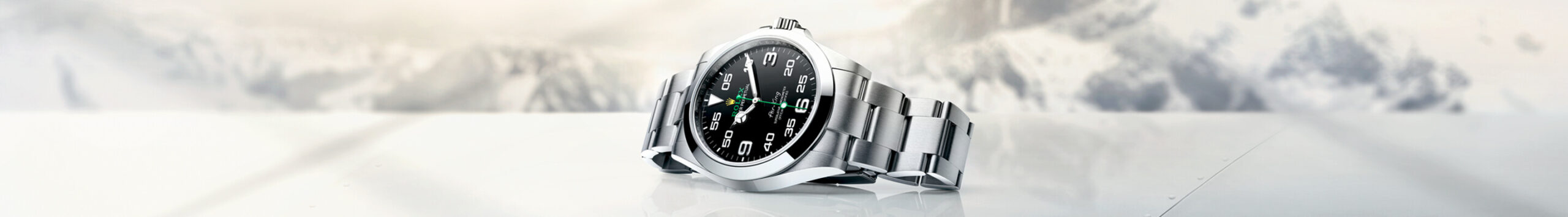 A silver analog wristwatch with a black dial and green hands on a reflective surface, set against a blurred winter landscape background.