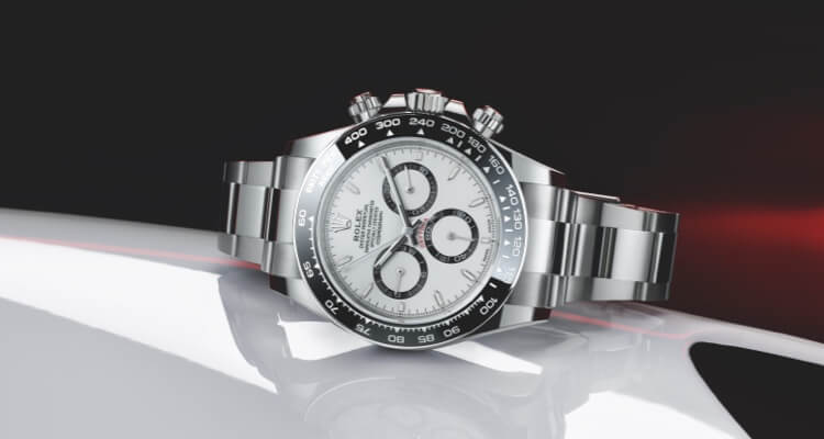 A stainless steel luxury watch with a white dial, three subdials, and a tachymeter bezel, displayed on a reflective surface with a red light accent.