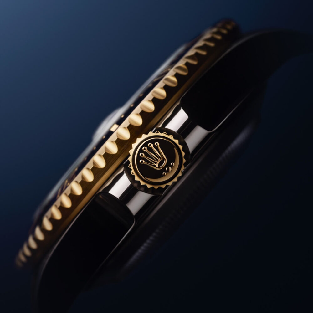 Close-up of a luxury watch crown and fluted bezel, displaying intricate details and a gold finish, against a dark blue background.