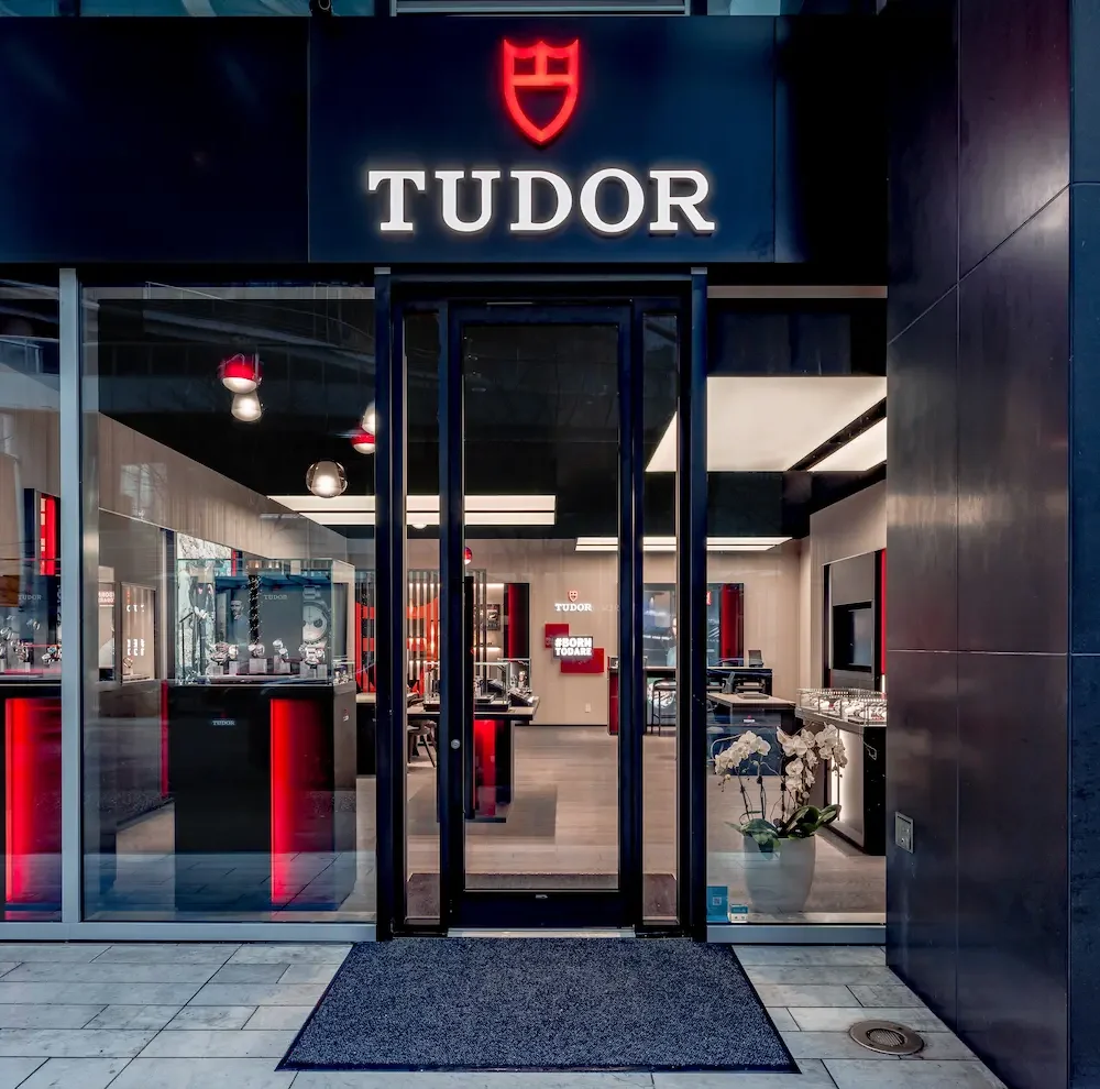 Front view of a Tudor watch store entrance with glass walls, displaying various watches and a red-accented modern interior.