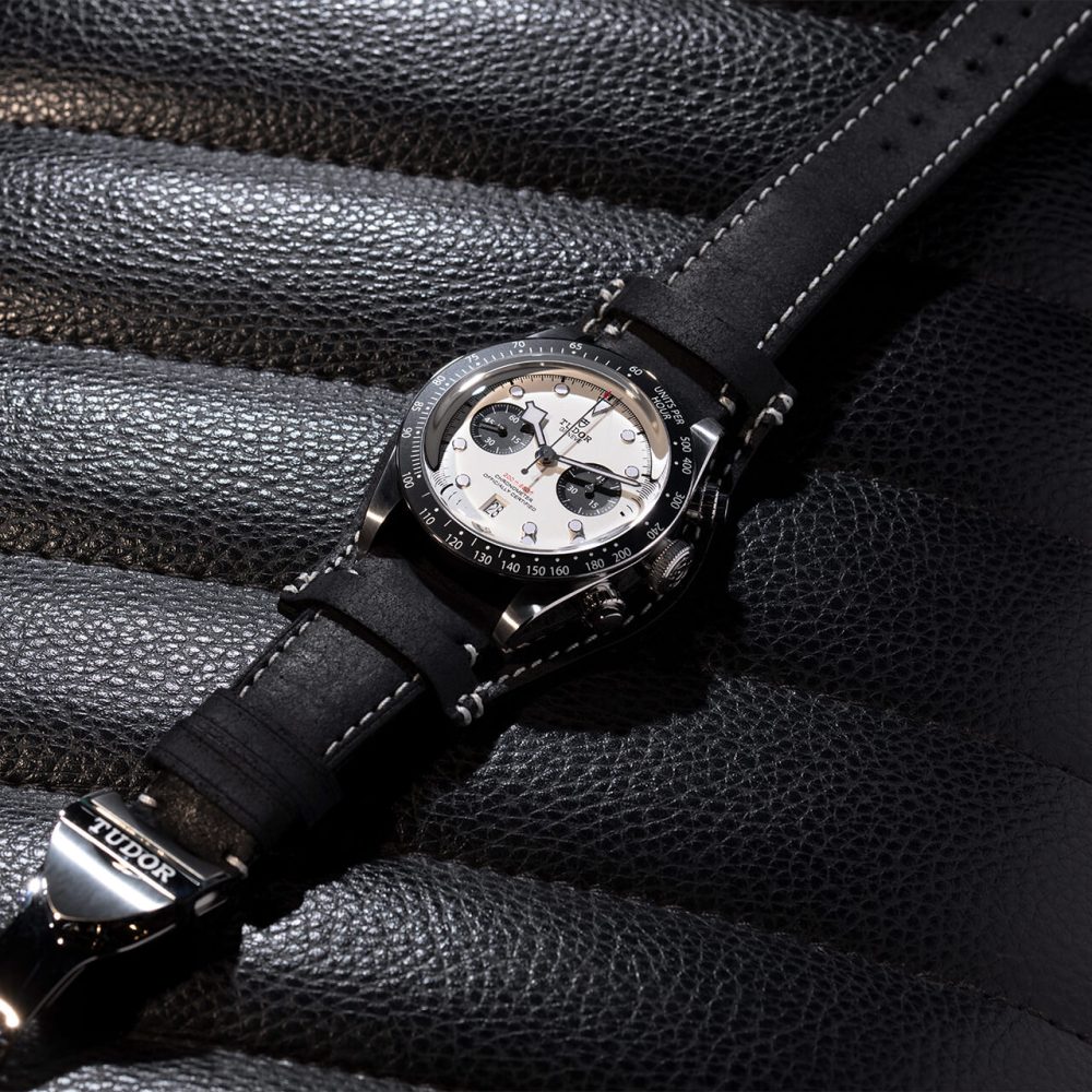 A luxury watch with a black leather strap resting on a textured surface, featuring a silver case and detailed dial with chronograph functions.